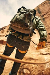 A climber wearing an olive green jacket, backpack, and yellow gloves rappels down a steep, rocky canyon wall.