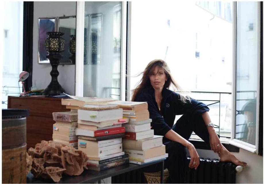 Woman sits on a radiator by a window in an apartment, surrounded by a stack of books on a table with decorative objects.