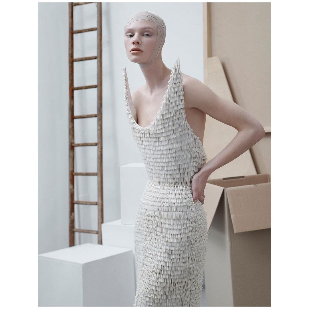 Female model in an off-white textured dress poses indoors with hands on hips, in front of boxes and a ladder. Fashion editorial.