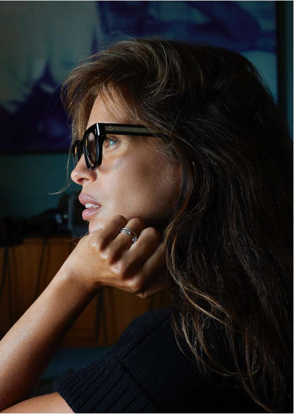 Close-up portrait of an adult woman wearing black-framed glasses, resting her chin on her hand, with long brown hair.