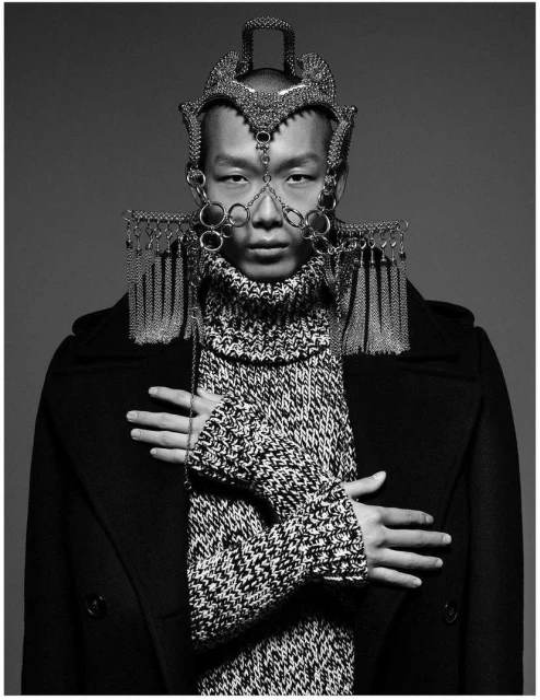 Monochrome studio portrait of a person wearing an ornate headpiece, turtleneck knitwear, and a dark overcoat against a gray backdrop.