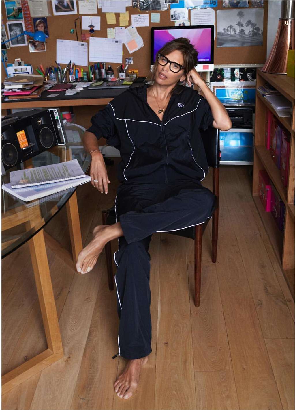 Adult woman in glasses and blue tracksuit sits barefoot indoors at a glass table, looking off camera with a relaxed expression.