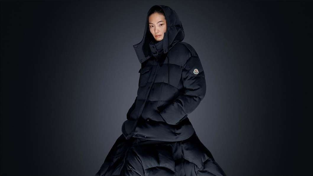 A woman wearing a Moncler black hooded puffer jacket and matching skirt poses against a dark gray studio backdrop for a fashion editorial.