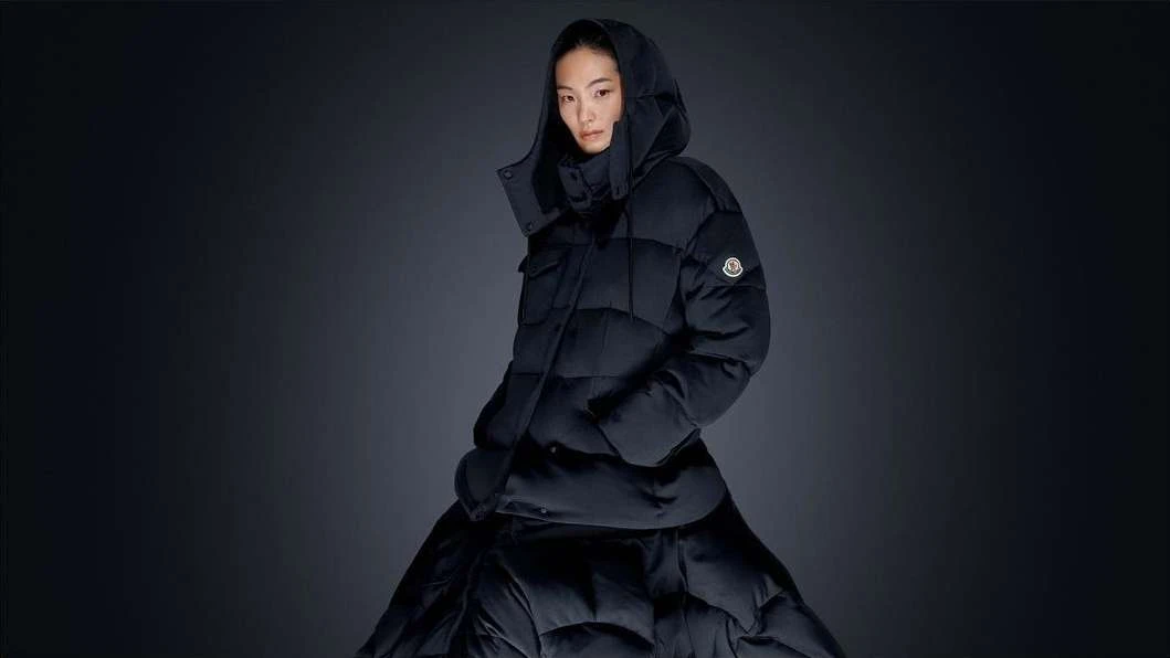 A woman wearing a Moncler black hooded puffer jacket and matching skirt poses against a dark gray studio backdrop for a fashion editorial.