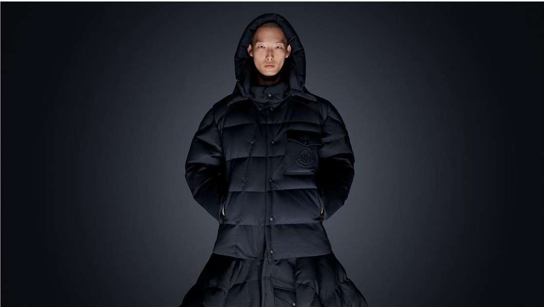 Person posing in a black puffer coat with a hood against a dark background, studio lighting highlights the jacket's details.