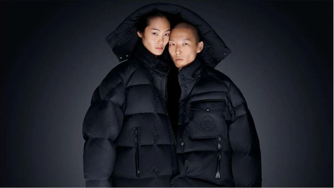 A stylish portrait features an Asian couple in hooded black puffer jackets against a dark backdrop, exuding a modern, fashionable aesthetic.