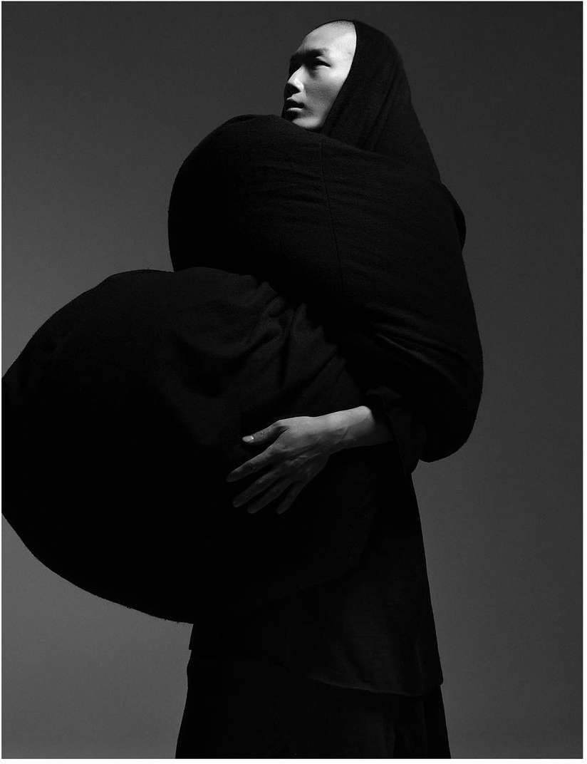 Monochrome studio portrait of an Asian male wearing a draped black fabric garment, his hands resting on his chest in a contemplative pose.