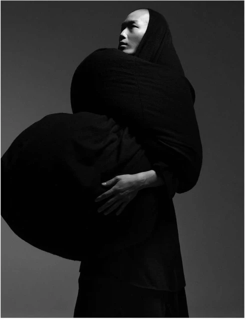 Monochrome studio portrait of an Asian male wearing a draped black fabric garment, his hands resting on his chest in a contemplative pose.