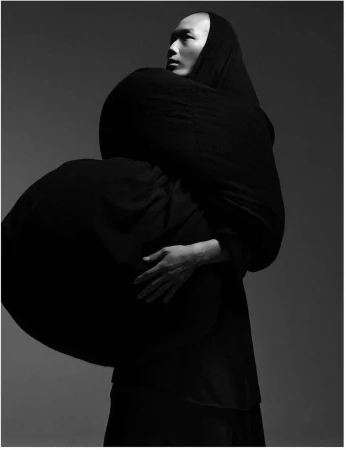 Monochrome studio portrait of an Asian male wearing a draped black fabric garment, his hands resting on his chest in a contemplative pose.