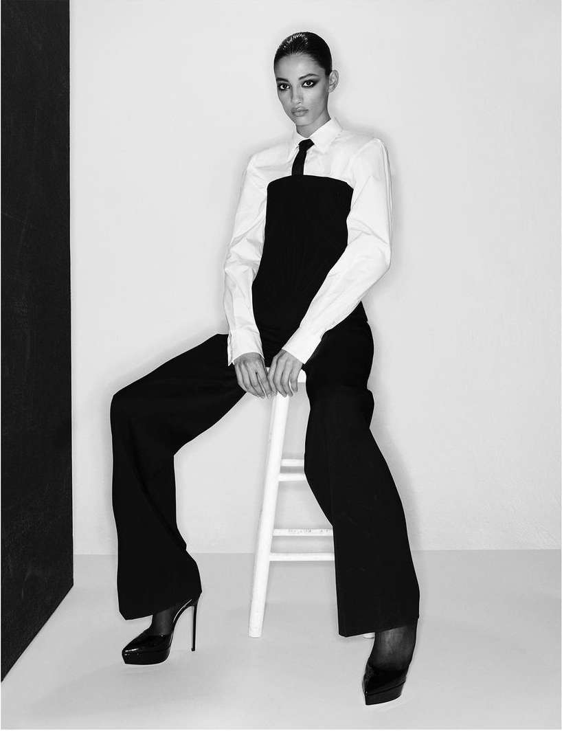 Elegant woman in black and white fashion sits on stool in a studio setting with soft lighting and sharp focus.