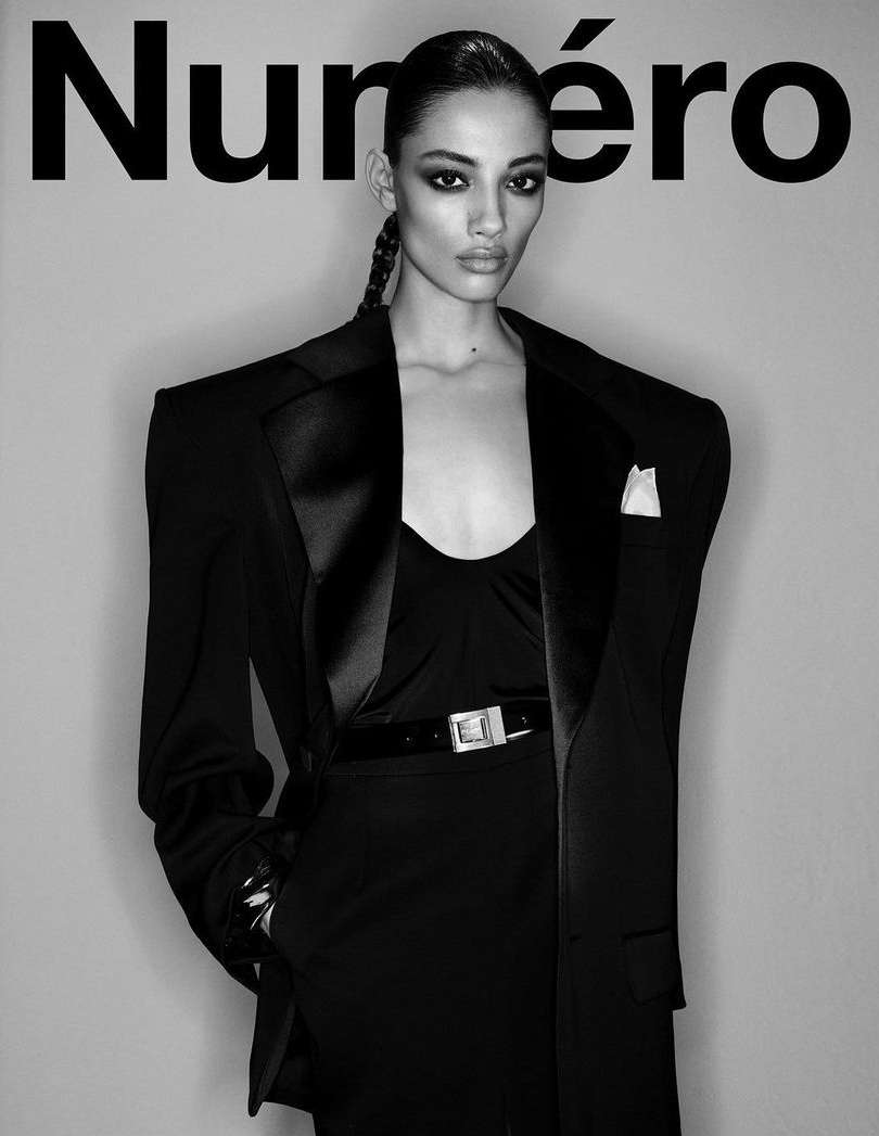 Fashion editorial portrait of a female model in an oversized black suit jacket and slicked-back hair against a grey backdrop.