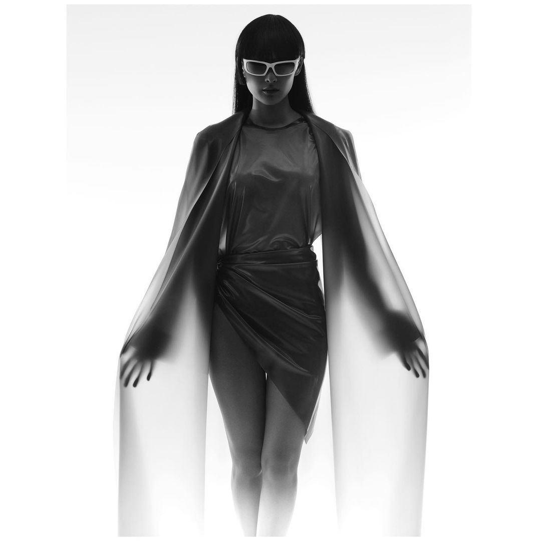 Monochrome studio shot of a woman wearing sunglasses, draped in sheer fabric, posing in front of a bright white background.