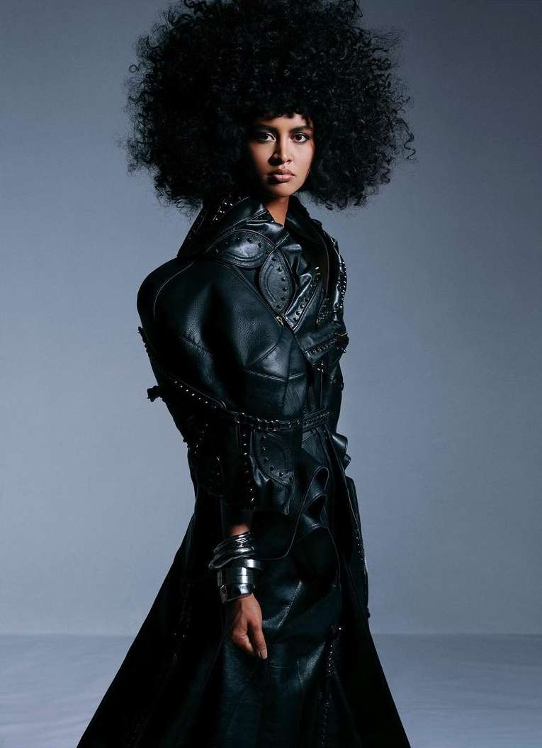 A striking fashion portrait of an adult female model with curly hair wearing a black leather trench coat against a grey studio backdrop.