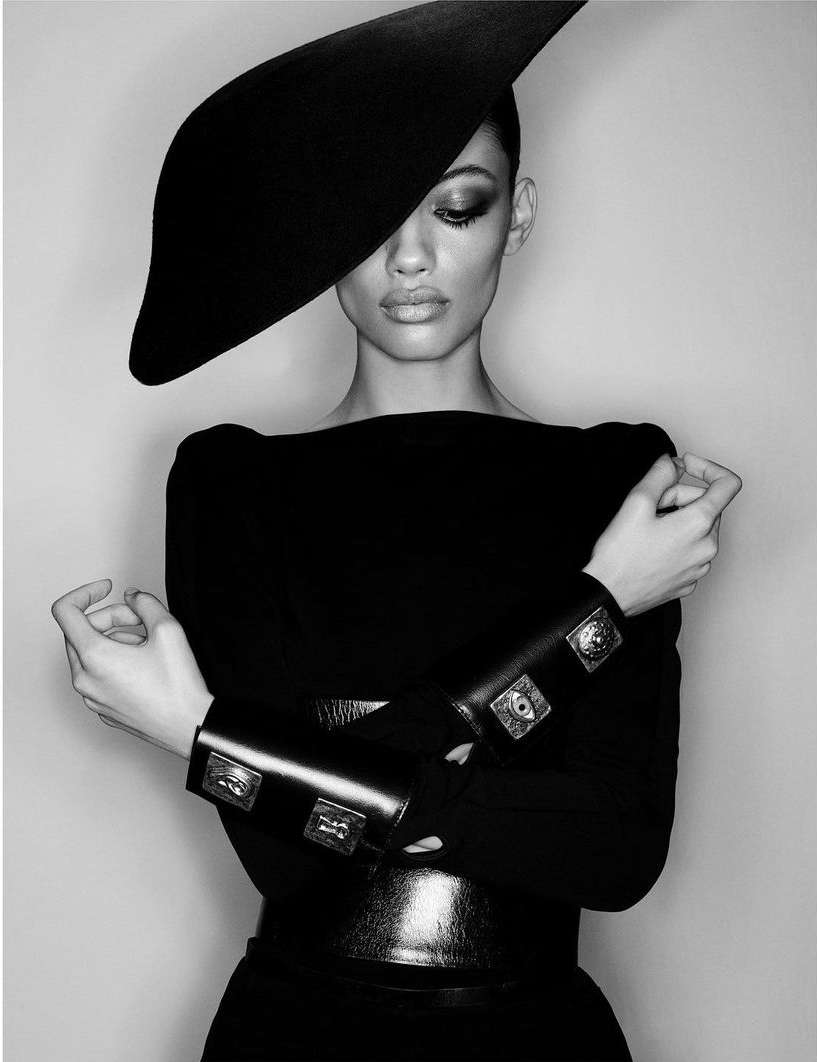 Black-and-white fashion shot: A woman wearing a black hat, dress, belt, and bracelet poses against a gray backdrop in a studio setting.