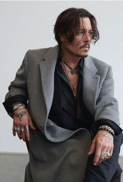 Portrait of Johnny Depp in a grey coat and black shirt, showcasing his unique style with rings and bracelets against a white background.