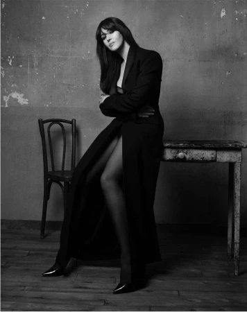 Monochrome portrait of Monica Bellucci seated on a rustic table in a long black coat against a weathered textured wall.