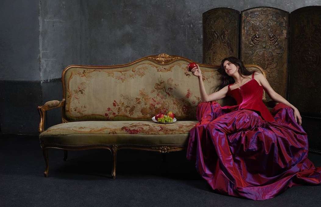 A woman lounges on an antique sofa, wearing a red gown, holding a red apple, in a dimly lit room, with a screen behind her.