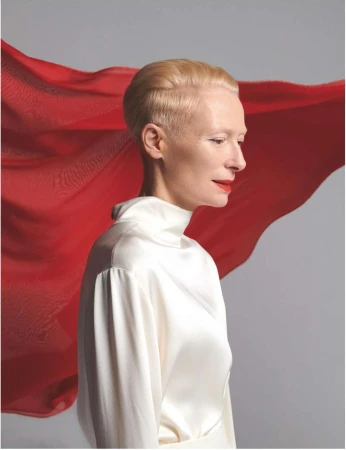 Tilda Swinton in a white high-neck silk top with a flowing red fabric behind her against a grey background.