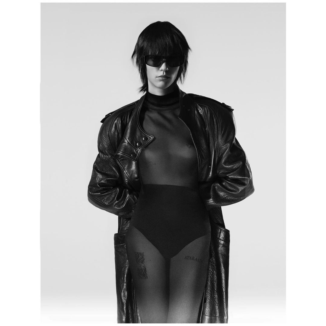 Monochrome studio shot of a woman with dark hair and sunglasses, wearing a black bodysuit and leather jacket against a white backdrop.