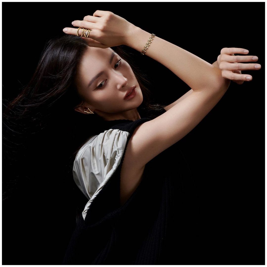 Woman in black dress with gold ring and bracelet on black background. Posing with hand near face, wearing jewelry. Fashion portrait.