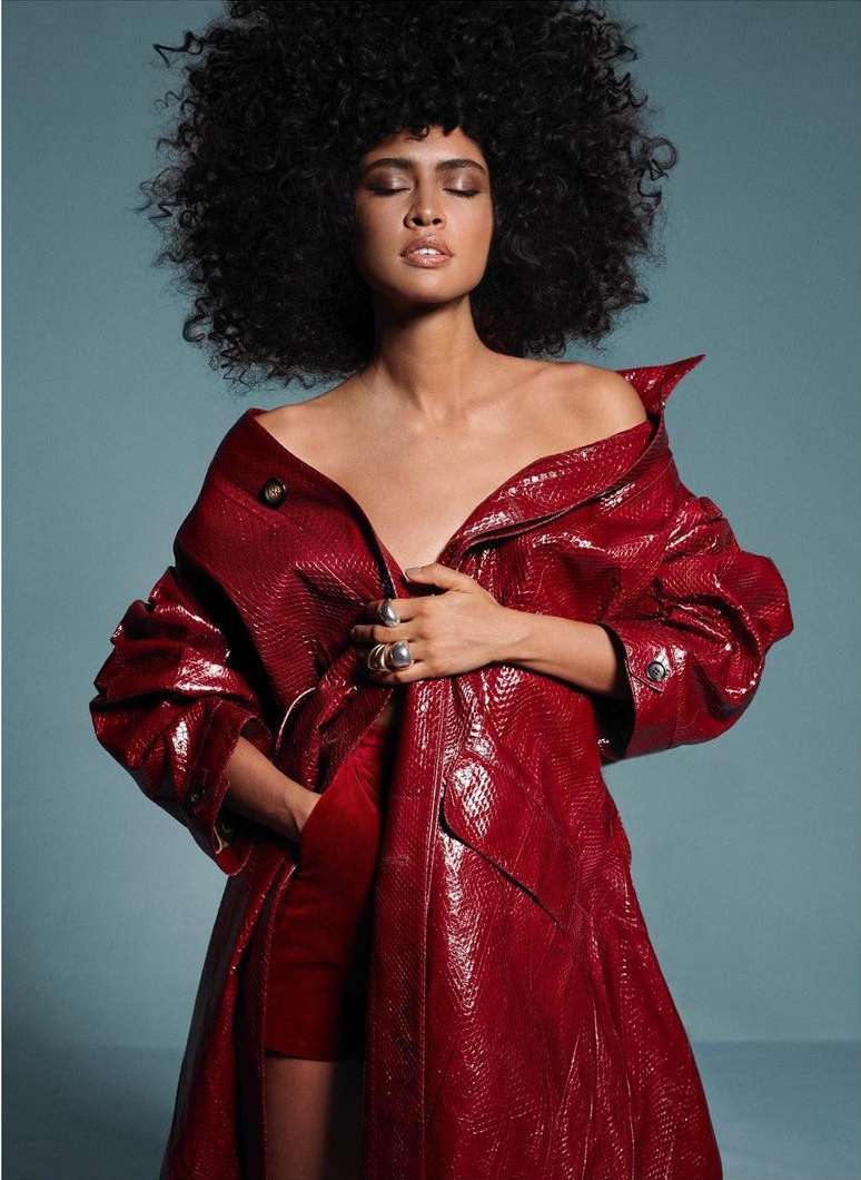 Glamorous woman in a glossy red coat poses against a blue backdrop. She has curly dark hair and silver rings on her fingers.