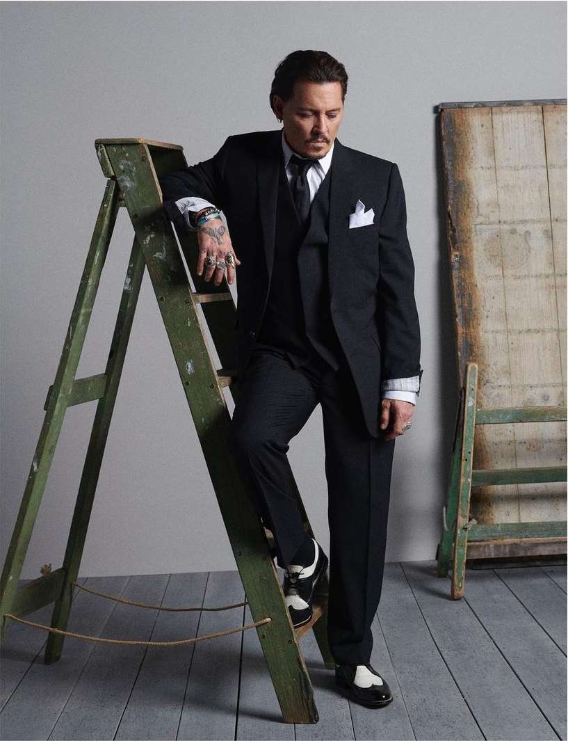 Johnny Depp poses in a dark suit next to a weathered green ladder in a studio. He's wearing black and white shoes.