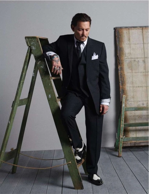 Johnny Depp poses in a dark suit next to a weathered green ladder in a studio. He's wearing black and white shoes.