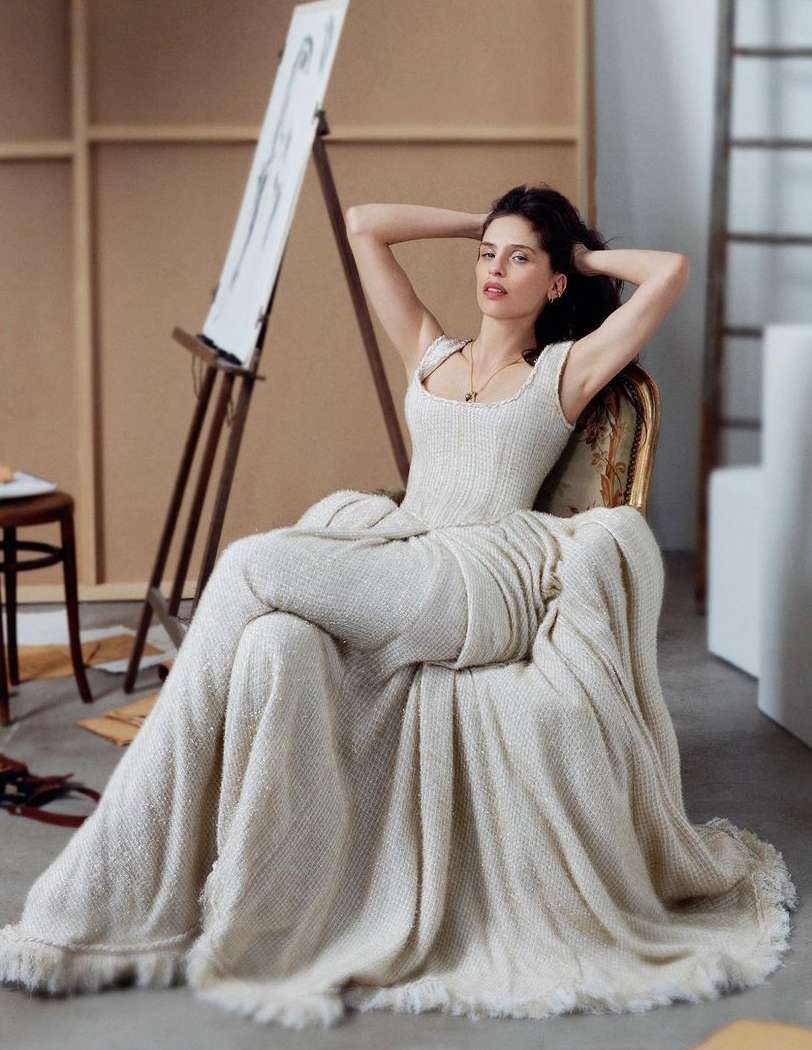 A woman in an off-white dress sits in an antique chair in front of an artist's easel and drawing in a well-lit studio.