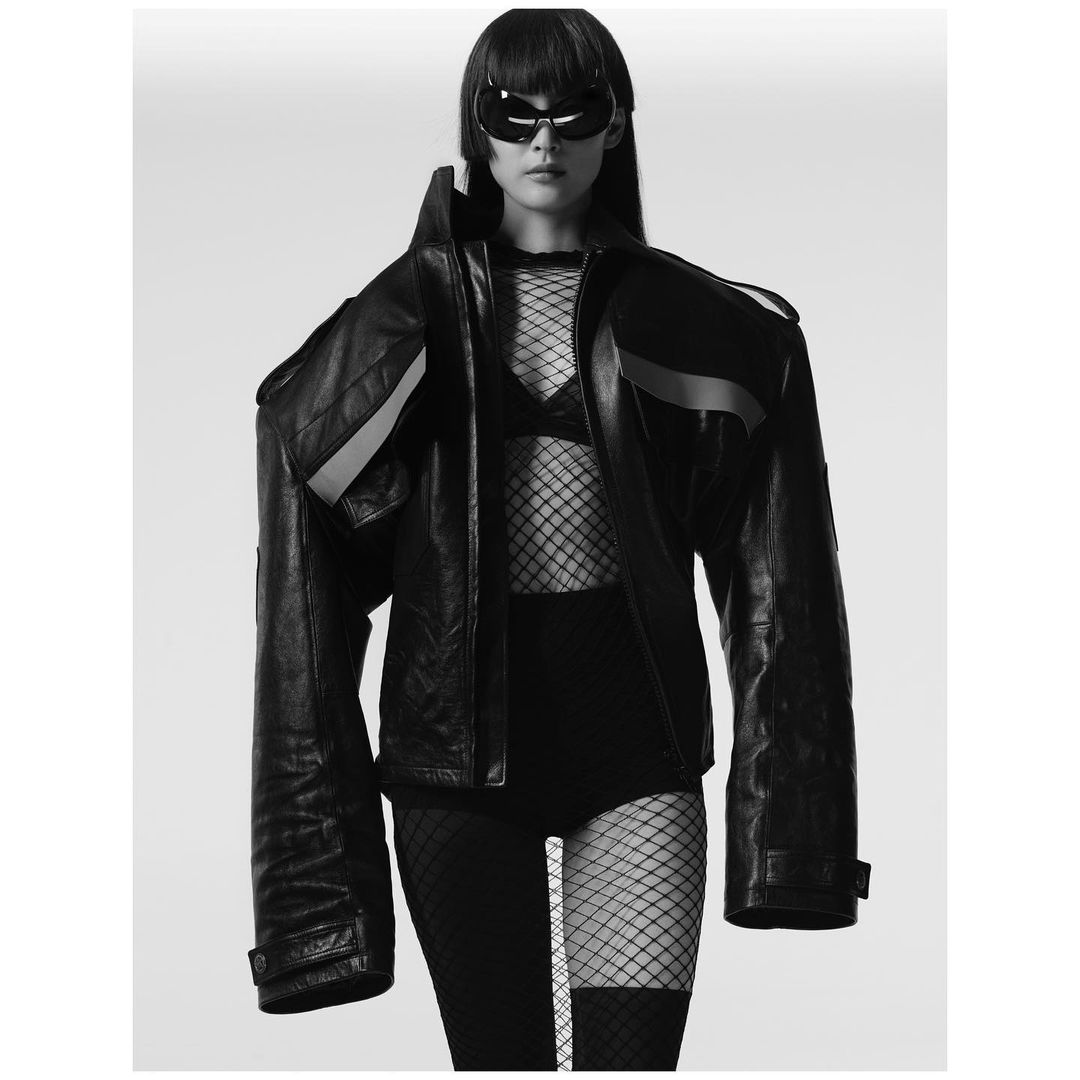 Black and white portrait of a female model in a leather jacket, bra top, sunglasses, and fishnet stockings against a white backdrop.