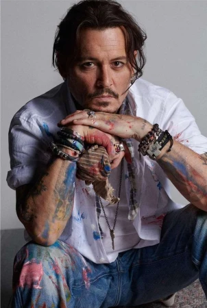 Portrait of Johnny Depp with paint-splattered shirt and jeans, hands clasped at chin, multiple bracelets, necklaces, and rings, neutral background.
