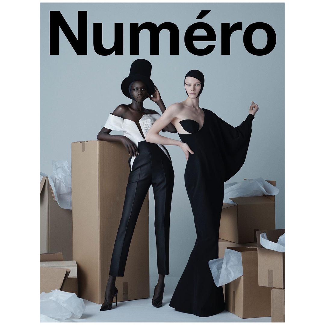 Two fashion models pose in black-and-white avant-garde outfits against a studio backdrop surrounded by cardboard boxes and packaging paper.