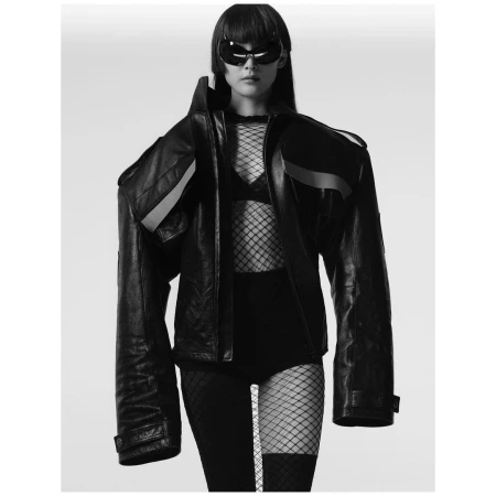 Model in oversized black leather jacket and futuristic sunglasses wearing fishnet bodysuit against a white backdrop.