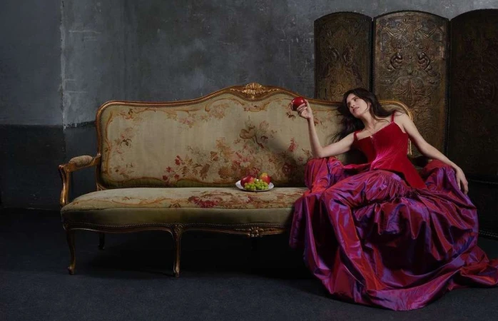 Woman in a red velvet top and purple silk skirt reclining on an ornate antique sofa, holding a red apple