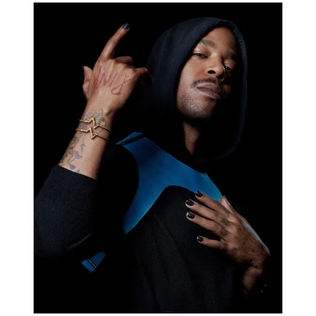 Portrait of a man in a black hoodie wearing a blue vest and gold bracelets against a black background