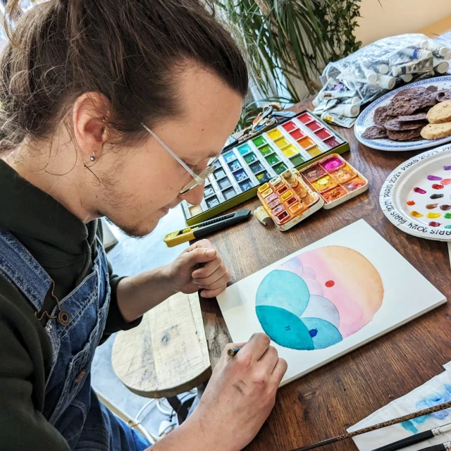 Adult male artist creating watercolor art at a wooden desk indoors, surrounded by paints, palette, and art supplies.