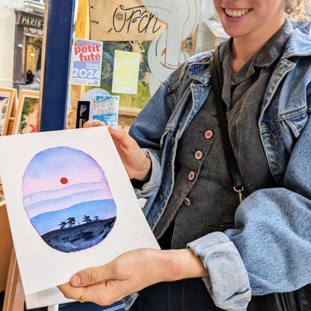 Person holding watercolor art depicting a landscape with a red sun, blue hills and dark trees on a table at an outdoor art fair.