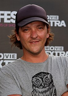 Headshot of adult male wearing a grey graphic t-shirt and grey baseball cap, smiling slightly, in front of a dark grey background.