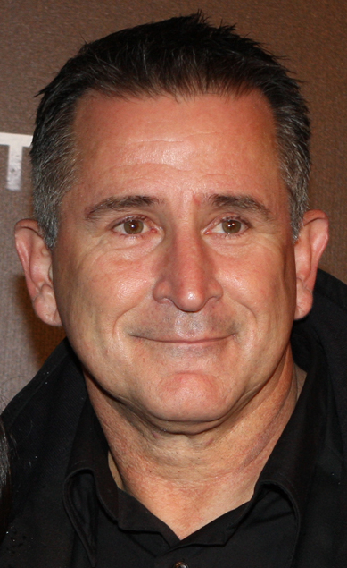 Portrait of mature adult male with short dark hair, fair skin, and a black collared shirt, posing against a dark brown background.