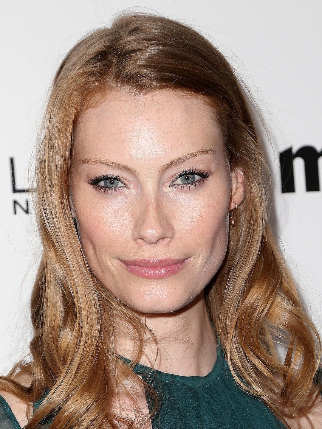 Close-up portrait of an adult female with wavy blonde hair and blue eyes, wearing a green dress. Makeup and soft lighting.