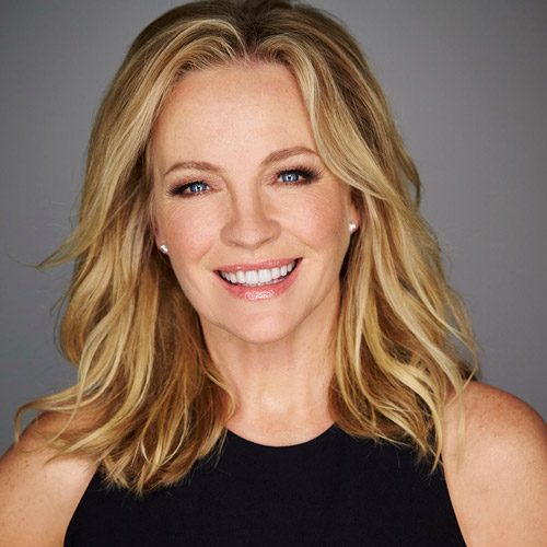 Portrait of a blond-haired, blue-eyed woman smiling in front of a solid grey background, wearing a black top and small pearl earrings.