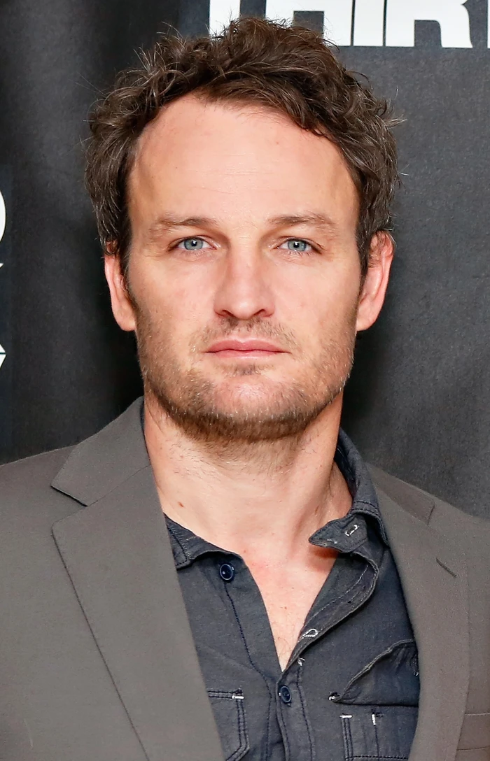 Portrait of an actor with short brown hair wearing a grey jacket and dark shirt against a dark backdrop