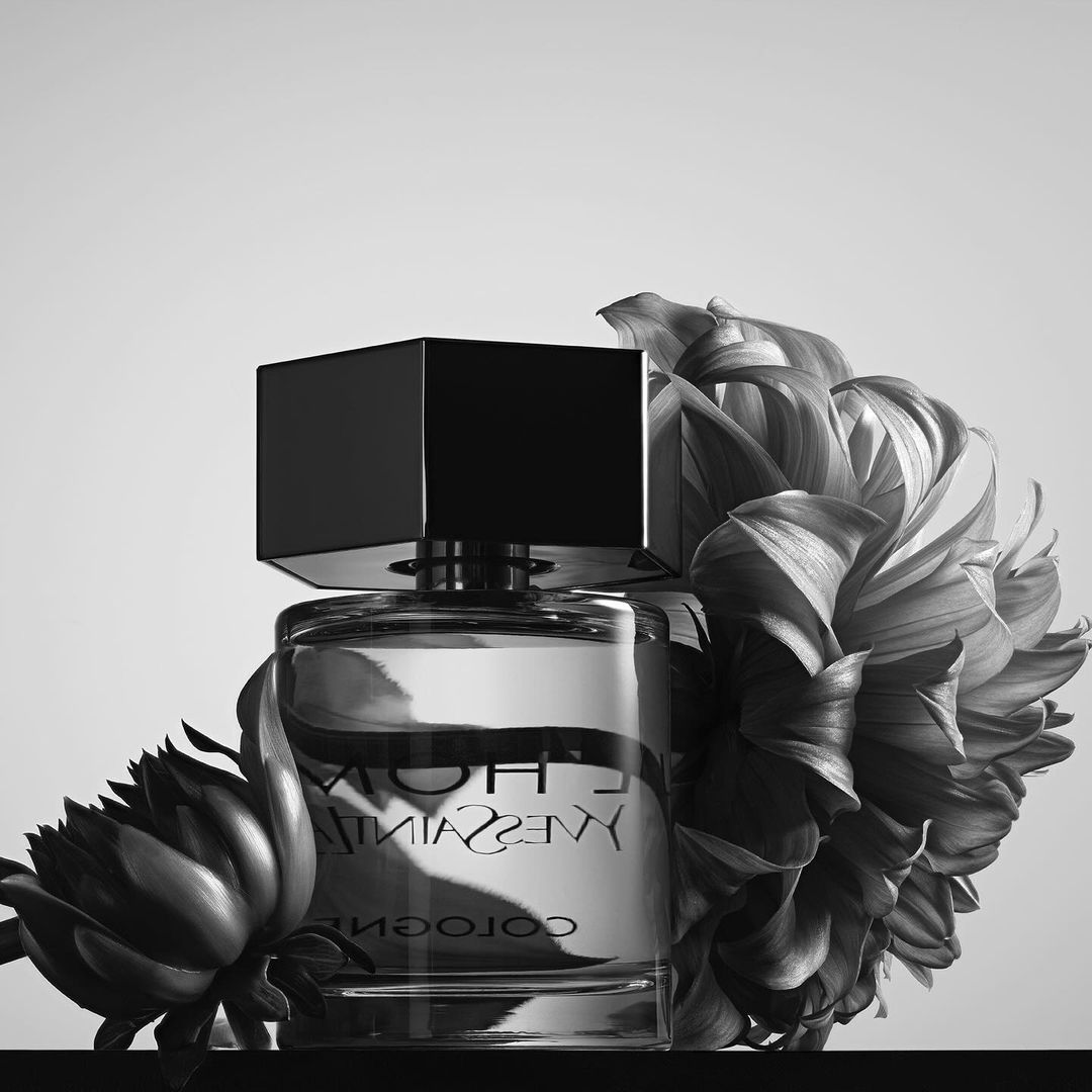 Black and white product photograph of a perfume bottle flanked by large draped flowers on a dark surface.