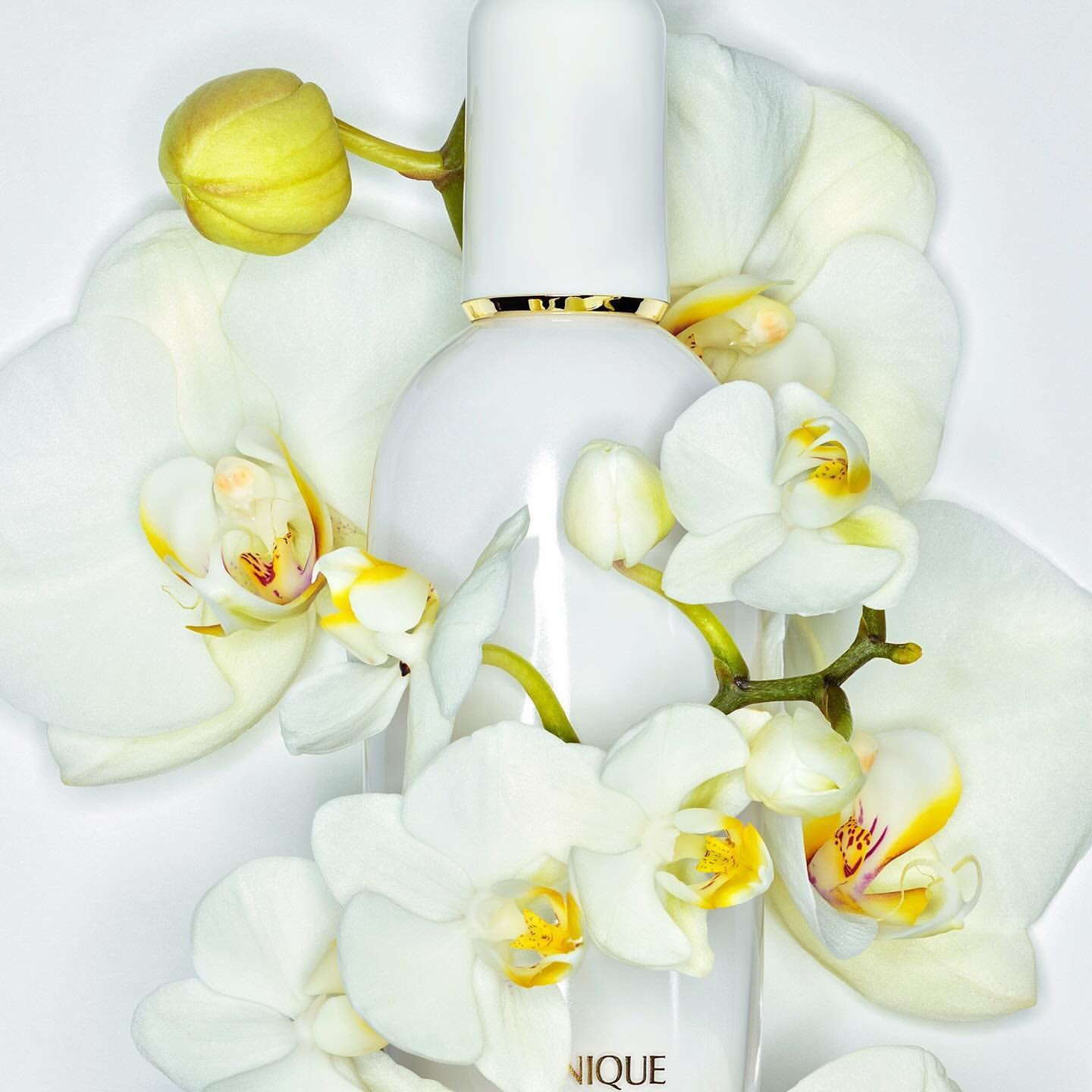 White skincare bottle surrounded by white orchid flowers, shot in a photo studio setting with bright lighting and soft shadows.