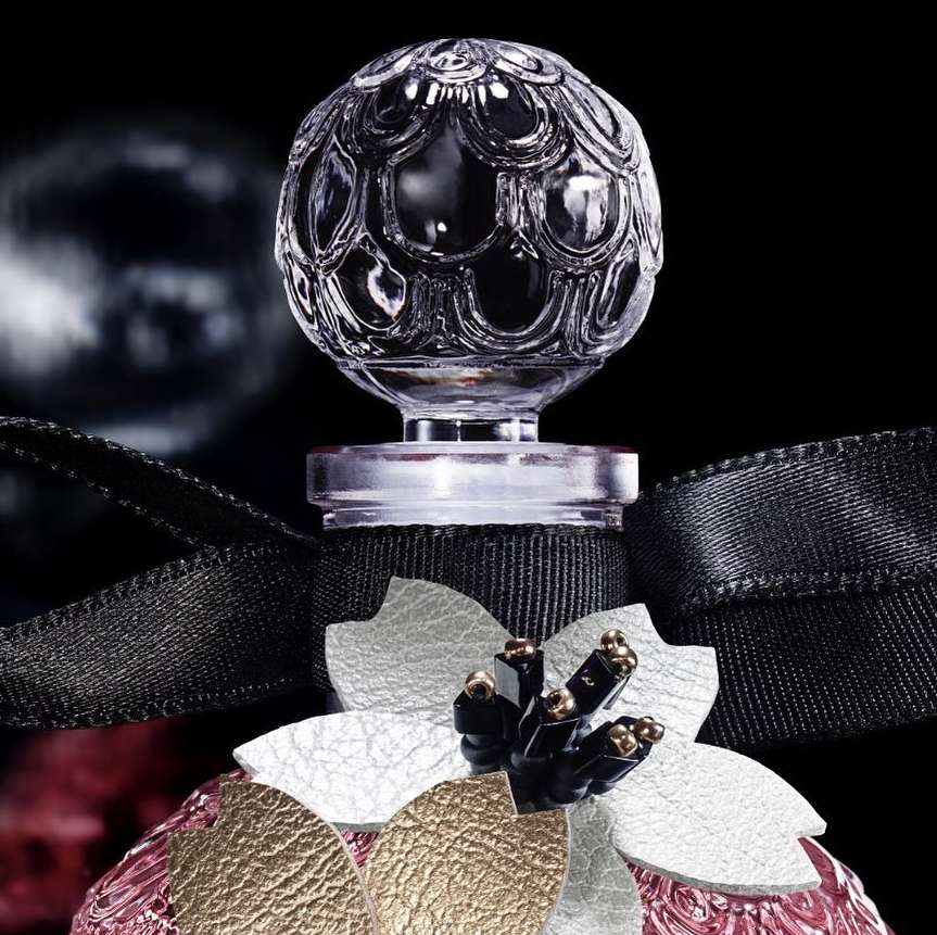 Close-up of a crystal perfume bottle stopper with ornate flower embellishments on a pink and gold textured bottle, set against a black background.