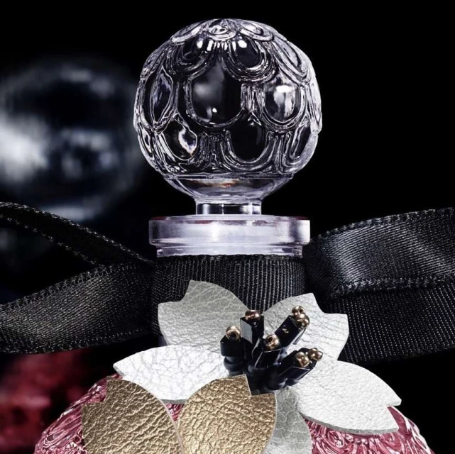 Close-up of a crystal perfume bottle stopper with ornate flower embellishments on a pink and gold textured bottle, set against a black background.