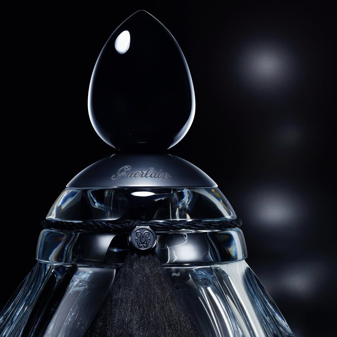 Elegant Guerlain perfume bottle with a black teardrop-shaped stopper, decorative cord, and a blurred background. High-end beauty product.