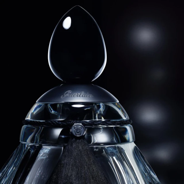 Elegant Guerlain perfume bottle with a black teardrop-shaped stopper, decorative cord, and a blurred background. High-end beauty product.