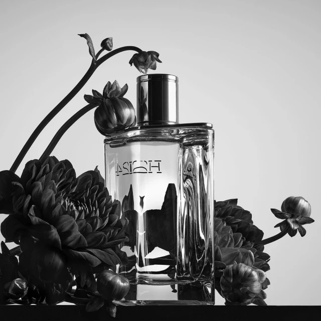 Monochrome product shot of clear glass perfume bottle with silver cap surrounded by dark floral arrangement against a white background.