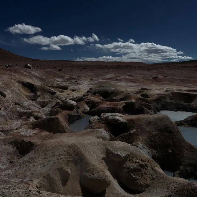 Desert landscape with brown rock formations, pools of water, and a dark blue sky with scattered white clouds on a sunny day.