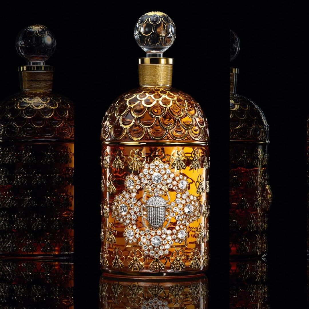 Guerlain perfume bottle with ornate, gold and jeweled design, featuring bees and a scarab, presented on a reflective black surface.